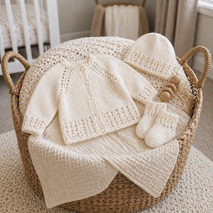 May include: A cream-coloured baby clothing set, including a cardigan, hat, and socks, arranged in a woven basket. The cardigan has button closures and decorative openwork. A wooden rattle is also included. The set is placed on a soft, cream-coloured blanket.
