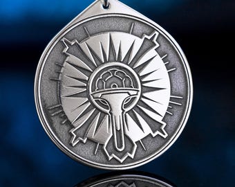 May include: Silver pendant with a detailed, circular design featuring a central emblem surrounded by radiating lines. The pendant hangs from a black cord. The design has a sunburst appearance, with a stylized symbol at the center.