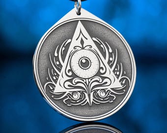 May include: A silver pendant with a black cord. The pendant features a detailed design of an eye within a triangle, surrounded by ornate patterns. The pendant is round with a triangular top, and is likely made of metal.