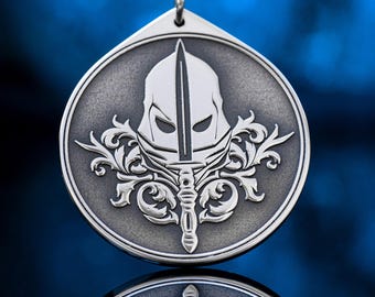 May include: Silver pendant with a detailed design of a helmet and sword, set against a circular backdrop with ornate flourishes. The pendant hangs from a black cord, showcasing a detailed, embossed design. The pendant is approximately 1.5 inches in diameter.