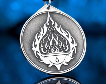 May include: Silver-toned pendant with a black cord. The pendant features a circular design with a flame motif. The flames are white and detailed, with a small drop shape at the bottom. The pendant is likely made of metal.