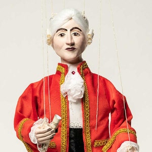 May include: A marionette puppet dressed in 18th-century attire. The puppet wears a red coat with gold trim, a white ruffled shirt, and a black vest. It has a powdered wig and holds a scroll. The puppet is suspended by strings.