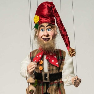 Puede incluir: Marioneta artesanal de un elfo sonriente con un largo gorro rojo, un chaleco a cuadros y una bufanda roja con lunares blancos. El elfo tiene una larga barba, ojos azules y sostiene una linterna de madera. La muñeca está suspendida por hilos.