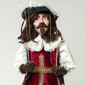 May include: A handcrafted marionette doll dressed in a historical costume. The doll features a black hat with a feather, brown hair, and a detailed face with blue eyes and a mustache. The doll's outfit includes a red vest with gold trim and a white ruffled collar.