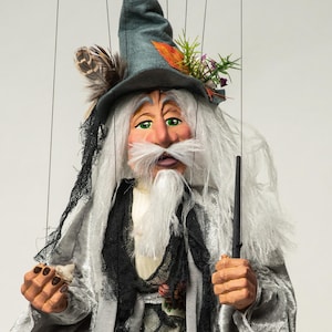 Puede incluir: Títere de mago artesanal con larga barba blanca, ojos verdes y sombrero gris adornado con plumas y flores. El títere viste una túnica plateada y sostiene una varita y un pequeño objeto.