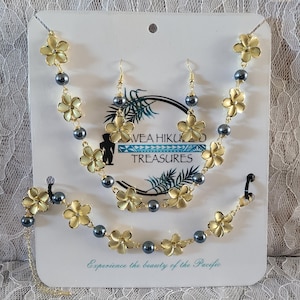 May include: A jewelry set featuring a necklace, bracelet, and earrings. The set includes gold-colored flower pendants and dark gray beads. The card behind the jewelry reads "AVEA HIKU TREASURES" and "Experience the beauty of the Pacific."