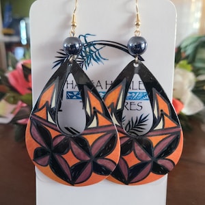 May include: A pair of teardrop-shaped earrings with a black border and a colorful floral design in orange, brown, and cream. Each earring has a dark gray bead and gold-colored hook. The earrings are displayed on a white card with text.