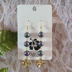 May include: A pair of dangle earrings with iridescent dark blue and purple beads and gold-colored floral charms. The earrings hang from gold-colored hooks. The earrings are displayed on a white card with a black floral design and the word "Aloha".