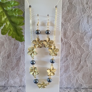 May include: A gold-toned necklace and earring set featuring flower-shaped pendants and dark gray beads. The necklace and earrings are displayed on a white card, with a lace background.