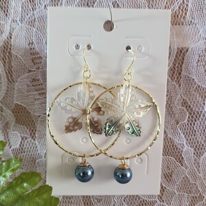 May include: Gold-tone hoop earrings with a leaf-shaped mother-of-pearl design and a dark blue bead. The earrings are displayed on a white card. The hoops are approximately 2 inches in diameter.