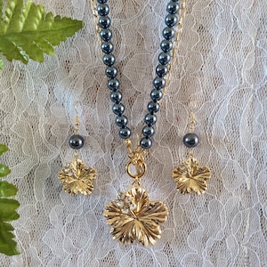 May include: A gold-toned necklace and earring set. The necklace features a strand of dark gray beads and a large gold flower pendant with clear rhinestones. The earrings have matching dark gray beads and gold flower charms. The jewelry is displayed on a white lace background.
