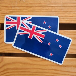 Può includere: Due adesivi rettangolari con la bandiera della Nuova Zelanda. Il design presenta la Union Jack nell'angolo in alto a sinistra, quattro stelle rosse e uno sfondo blu navy. Gli adesivi sono su una superficie di legno.