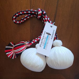 May include: A pair of white, round, fabric-covered objects with a braided black, red, and white cord. A tag reads "Have a Hikuleo Treasures" and "Handmade by". The cord is tied with a red, black, and white tassel.