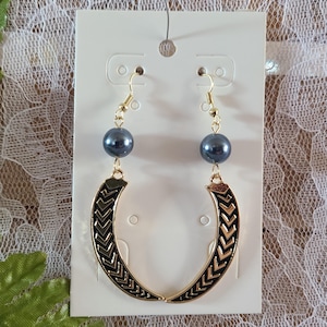 Può includere: Un paio di orecchini color oro con una perla grigio scuro e un design a forma di mezzaluna nero e oro. Gli orecchini sono appesi a una carta bianca con uno sfondo di pizzo.
