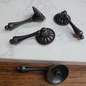 May include: Four antique bronze drawer pulls with a decorative design. Each pull features a round backplate with a ridged pattern, a curved handle, and a small ball at the end. The pulls are arranged on a white and brown surface.