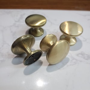 May include: A collection of five brass cabinet knobs. Each knob features a rounded top and a flared base, with a screw attachment on the underside. The knobs have a warm, golden hue and a smooth, polished finish. Ideal for furniture upgrades.