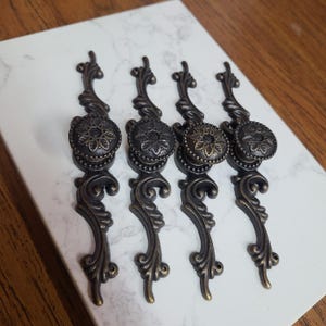 May include: Four antique bronze cabinet pulls with ornate floral and scroll designs. Each pull features a round knob with a detailed flower pattern and a decorative base. The pulls are arranged on a white marble-like surface.