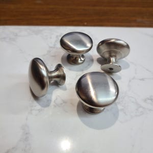 May include: Four brushed nickel cabinet knobs with a rounded design. The knobs have a circular top and a short, cylindrical base. They are arranged on a white and gray marble-patterned surface.