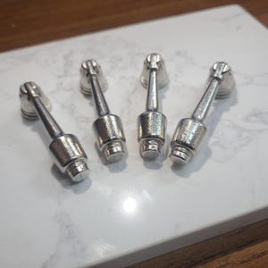 May include: Four silver-colored metal objects with a cylindrical body and a decorative top. The objects are arranged on a white and gray marble surface. The objects appear to be decorative or functional hardware.