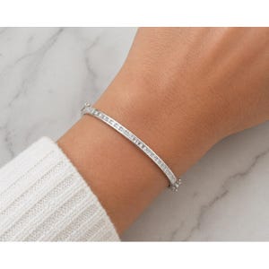 May include: A silver bangle bracelet with a row of small, clear stones set along the top. The bracelet is worn on a wrist, and the background is a marble-like surface.