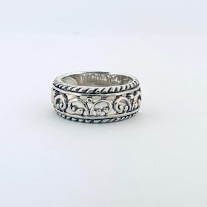 Puede incluir: Un anillo de plata con un diseño calado ornamentado. El anillo presenta un patrón repetitivo de formas arremolinadas y bordes con cuentas. El anillo está sobre un fondo blanco.