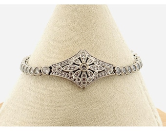 May include: Silver-toned bracelet featuring a central diamond-shaped design with a floral motif, surrounded by small, round, clear stones. The bracelet is displayed on a beige surface, showcasing its intricate details and elegant style.