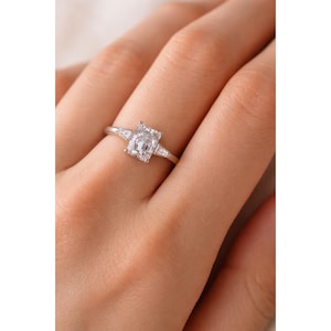 May include: A silver ring with a large, rectangular-cut clear gemstone as the focal point, with two smaller, triangular-cut stones on either side. The ring is set on a slender band, displaying a classic and elegant design. The ring is shown on a finger.