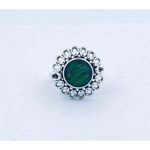May include: A silver-toned ring featuring a round, green stone with a swirling pattern. The stone is surrounded by a halo of small, open circles, creating a floral-like design. The ring is set against a white background.