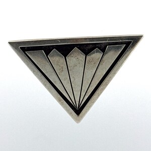 May include: A silver-coloured triangular brooch with a geometric design. The brooch has a black border and radiating lines, creating a fan-like pattern. The brooch is set against a white background.