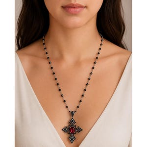 May include: A necklace featuring a silver-toned cross pendant with a central oval red gemstone and black accents, suspended from a delicate chain adorned with small, round black beads that are evenly spaced along the entire length of the metal chain.