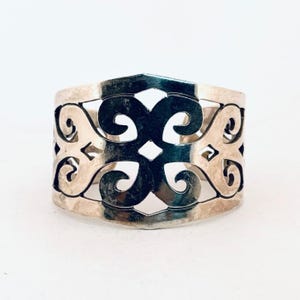 May include: A silver-toned cuff bracelet with an openwork design. The bracelet features a repeating pattern of swirling shapes and geometric cutouts. The metal has a polished finish, reflecting light. The cuff is wide, approximately 2 inches in width.