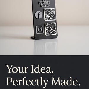 May include: A black display stand with the text "GUIDED BY ANGELS" and social media icons for Facebook and Instagram, each with a corresponding QR code. The stand also features the text "Your Idea, Perfectly Made."