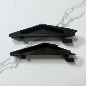 May include: Two black, triangular shelf brackets are shown against a white and grey marble-effect surface. The brackets have a shelf and a curved support. The top bracket is smooth, while the bottom one has a textured surface.