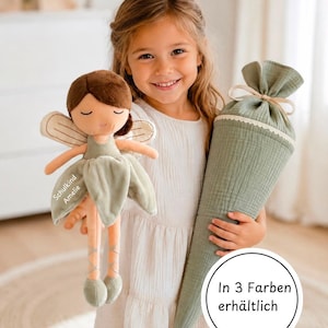 Könnte beinhalten: Eine Plüsch-Feenpuppe mit braunen Haaren, geschlossenen Augen und einem salbeigrünen Kleid, das den Text 'Schulkind Amelie' auf dem Rock trägt. Daneben befindet sich eine salbeigrüne Stofftüte mit Schleife und Spitzenborte sowie ein kreisförmiges Etikett mit der Aufschrift 'In 3 Farben erhältlich'.