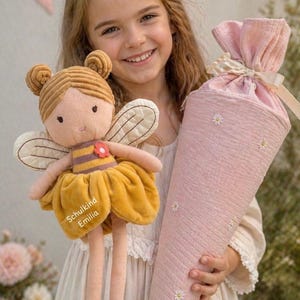 Könnte beinhalten: Eine Plüsch-Bienenpuppe mit braunen Haaren, gestreiftem Oberteil und gelbem Rock mit der Aufschrift Schulkind Emilia, neben einer kegelförmigen rosa Geschenktüte aus Stoff, verziert mit kleinen weißen gestickten Gänseblümchen und einer Schleife.
