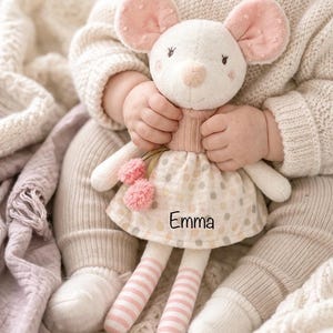 Könnte beinhalten: Eine weiche, plüschige Mauspuppe mit rosa Ohren, einer rosa Nase und einem gepunkteten Kleid. Die Puppe hat gestreifte Beine und zwei rosa Pompons. Der Name "Emma" ist auf das Kleid gedruckt. Die Puppe wird von einem Baby gehalten.