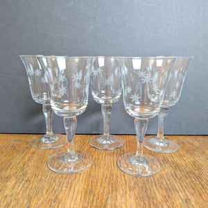 May include: Five clear glass wine glasses with etched starburst designs. The glasses have a classic stem and a wide, rounded bowl. They are arranged on a wooden surface against a dark gray background.