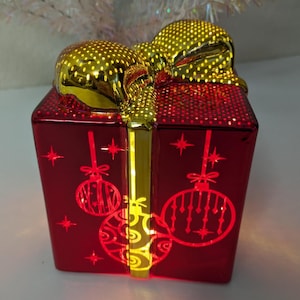 May include: A red, illuminated gift box with a gold bow. The box features cut-out Christmas ornament designs and star accents. The bow and ribbon have a gold, polka-dot pattern. A festive holiday decoration.