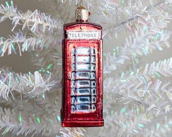 May include: A red glass Christmas ornament shaped like a British telephone booth. The ornament has the word "TELEPHONE" at the top and is hanging on a white Christmas tree. The ornament is approximately 4 inches tall.