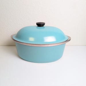 May include: A round, light blue casserole dish with a matching domed lid. The lid features a small, black, circular knob handle at the center. The pot has a smooth, solid-colored finish and a metallic rim visible between the base and the lid.