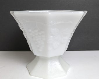 May include: A white, footed, hexagonal-shaped bowl. The bowl features a grape and leaf design on the sides. The bowl is made of a milky white glass and has a textured surface. The bowl is likely vintage and suitable for serving.