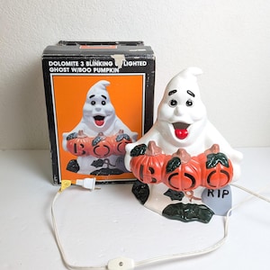 May include: A white ceramic Halloween ghost figurine holding three orange pumpkins with the word "BOO" on them. The ghost has a red tongue and black eyes. The box behind the figurine reads "DOLOMITE 3 BLINKING LIGHTED GHOST W/BOO PUMPKIN".
