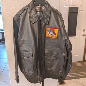 May include: A dark brown leather jacket with a USMC Devil Dog patch. The jacket has two front pockets, a zipper closure, and a collar. The patch features a bulldog wearing a cowboy hat and the text "USMC DEVIL DOG".