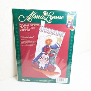 May include: A packaged cross-stitch kit for a holiday stocking. The kit features a design of an angel and a child, with the name "SAMANTHA" embroidered on the stocking. The package is green and red, with the brand name "Alma Lynne". The stocking is 18 inches (45.7cm) diagonal.
