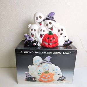 May include: A ceramic Halloween night light featuring smiling ghosts, a red jack-o'-lantern, and a black cat wearing a witch hat. The light sits atop a box that reads "BLINKING HALLOWEEN NIGHT LIGHT". The ghosts wear purple witch hats and boots.