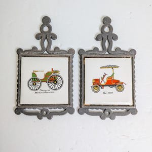 May include: Two vintage-style decorative wall hangings. Each features a square white tile with a colorful illustration of an antique car. The tiles are set in ornate, gray metal frames. One reads "Duryea's Motorwagon 1895" and the other "Reo - 1905".