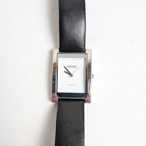 May include: A rectangular Gucci quartz watch with a silver-tone case and a black leather band. The white watch face displays the Gucci logo and the word "QUARTZ". The watch has silver hands and a silver crown.