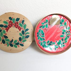 May include: Two round Christmas-themed items. One is a wooden lid with a wreath design, featuring green leaves, red berries, and a plaid bow. The other is a red plate with a similar wreath design, packaged in a cardboard box.