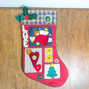 May include: Red Christmas stocking with patchwork designs. Features an angel, hearts, stars, and a Christmas tree. The top is plaid with a green holly and berry accent. A festive holiday decoration.