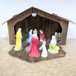 May include: A nativity scene set featuring a wooden-style stable structure and multiple small figurines. The figures include individuals in bright yellow, red, blue, and green robes, along with white animal figures, all arranged on a base.